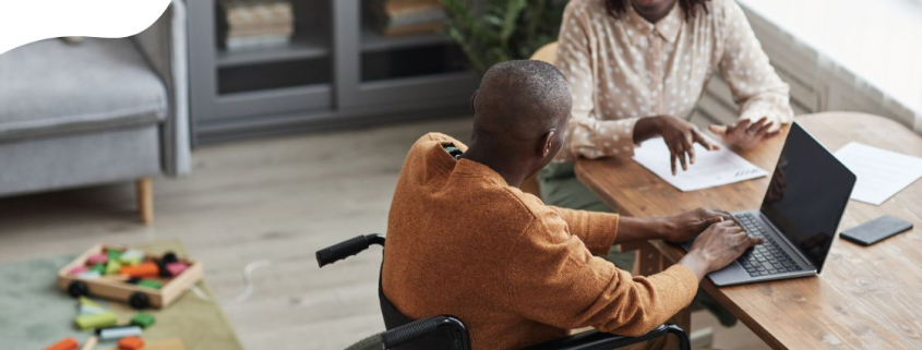 homem cadeirante com uma instrutora o auxiliando no computador. Ambos possuem a cor da pela negra e estão conversando com um computador na mesa sobre acessibilidade digital.